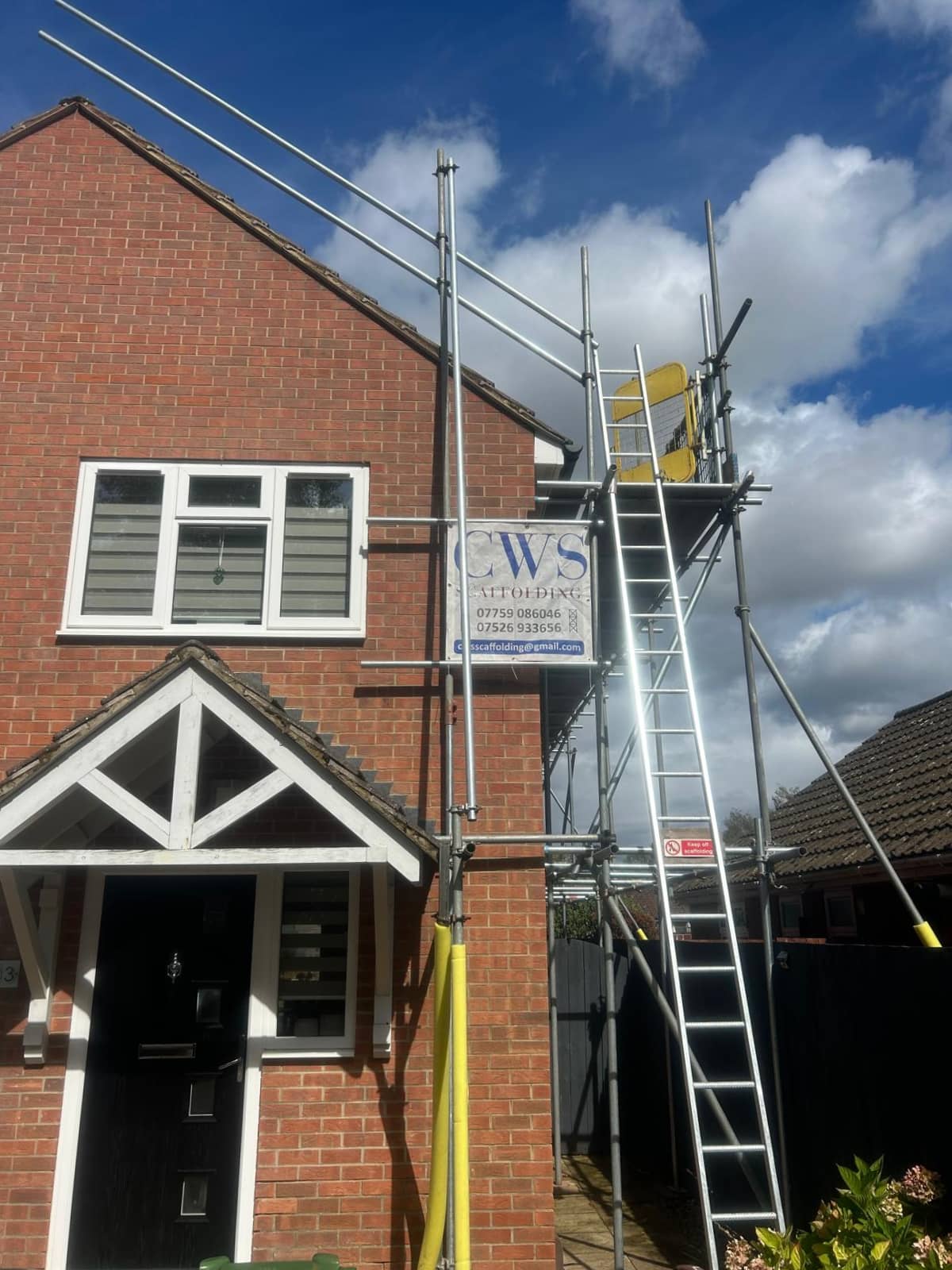 A brick house with scaffolding set up along one side, a ladder leaning on the structure, and a sign reading "CWS Attwood" attached to the scaffolding. The sky is partly cloudy.