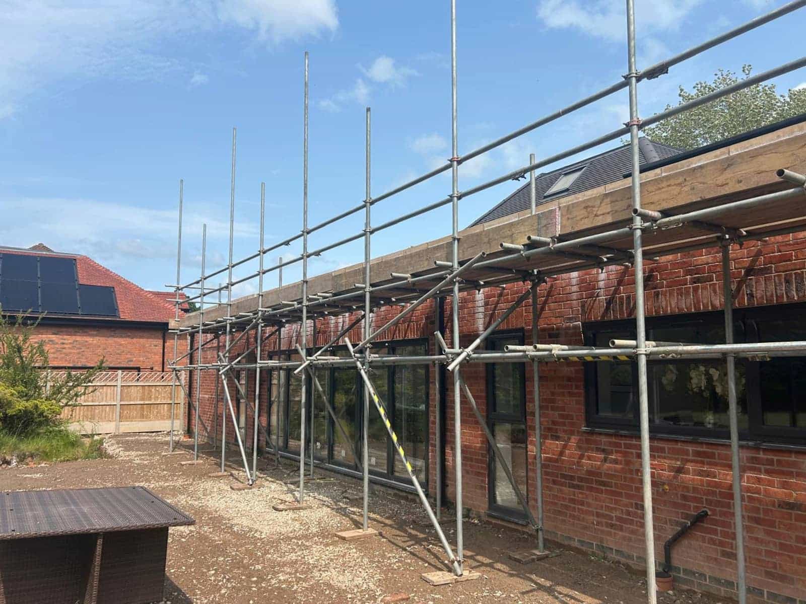 Scaffolding is set up along the brick exterior of a building with several windows. The sky is clear, and there is a wooden fence, a table, and another building in the background.