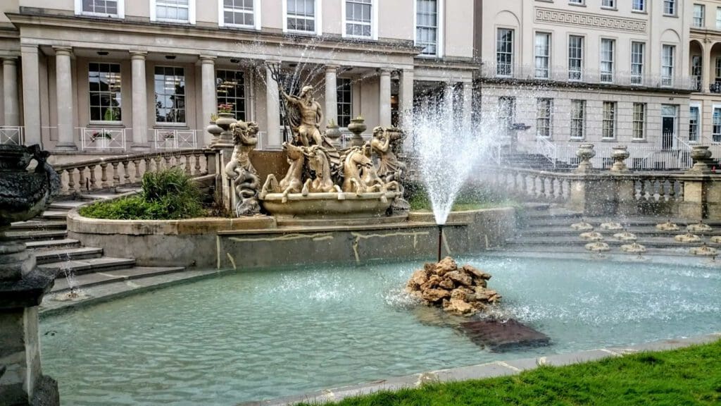 A decorative fountain with sculptures of figures and horses stands in front of a grand, historic building with tall windows and columns. Water sprays upward from the fountain into a shallow pool.