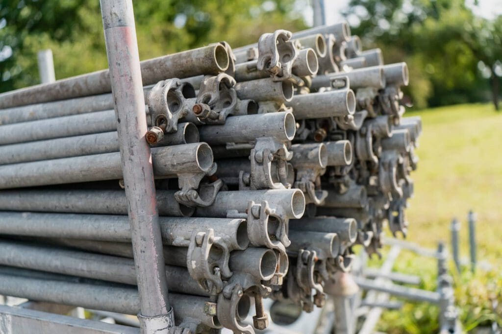A stack of metal scaffold pipes with clamps is arranged horizontally outdoors on a sunny day, with green grass and trees visible in the background.