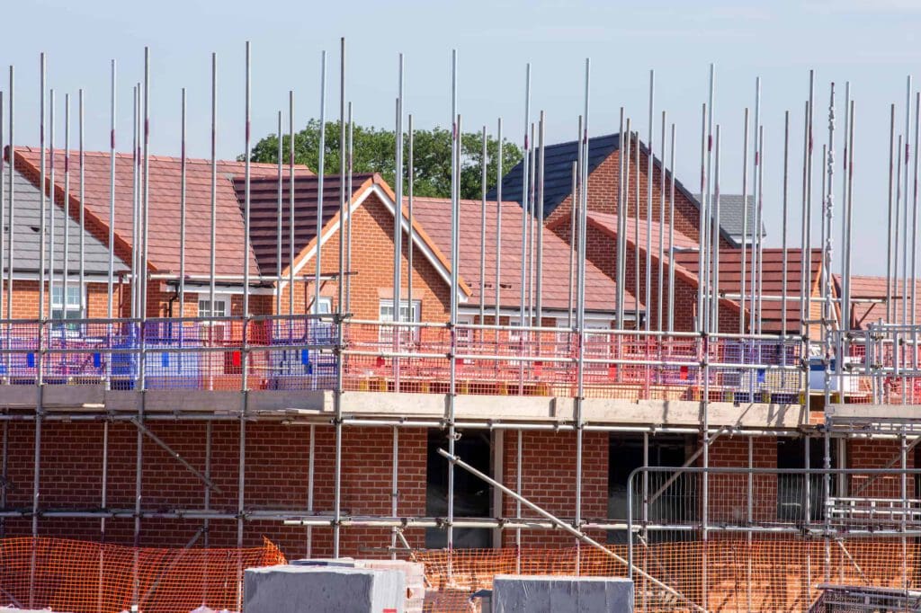 Rows of red-brick houses under construction, with scaffolding, metal poles, and safety barriers in place. Blue sky and green trees are visible in the background.