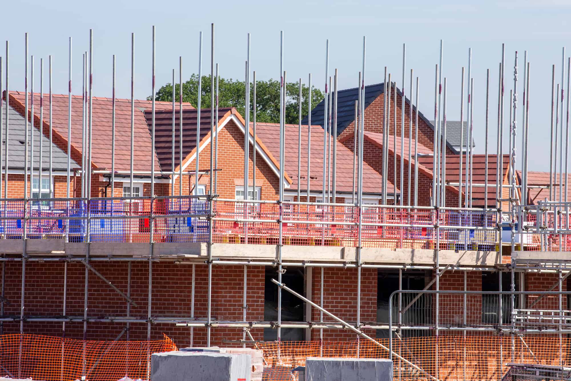 Rows of red-brick houses under construction, with scaffolding, metal poles, and safety barriers in place. Blue sky and green trees are visible in the background.