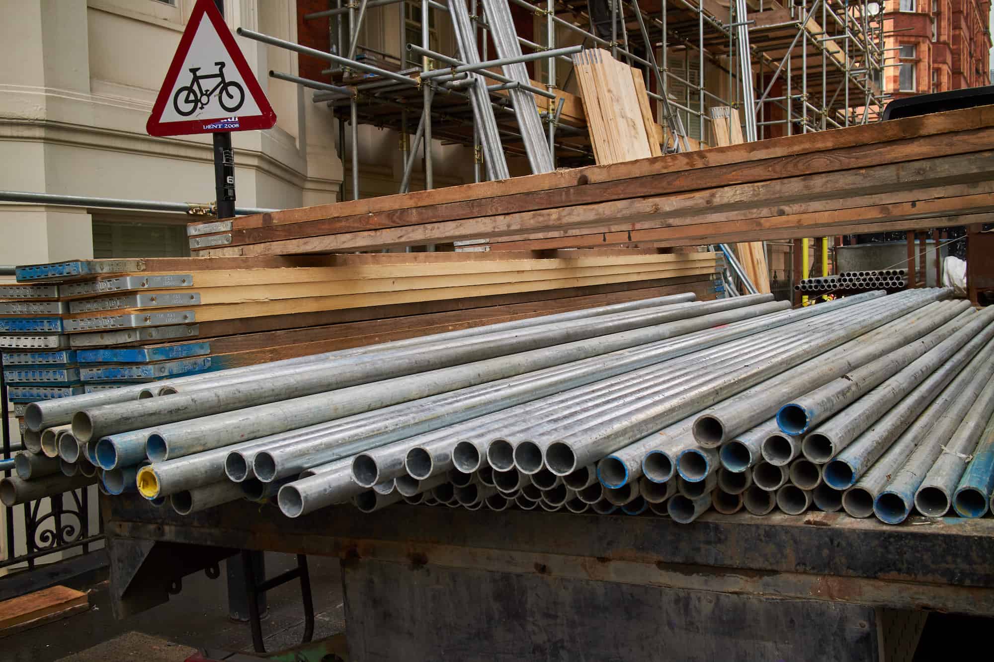 Stacked metal pipes and wooden planks are loaded onto a trailer at a construction site, with scaffolding and a bicycle crossing warning sign visible in the background.