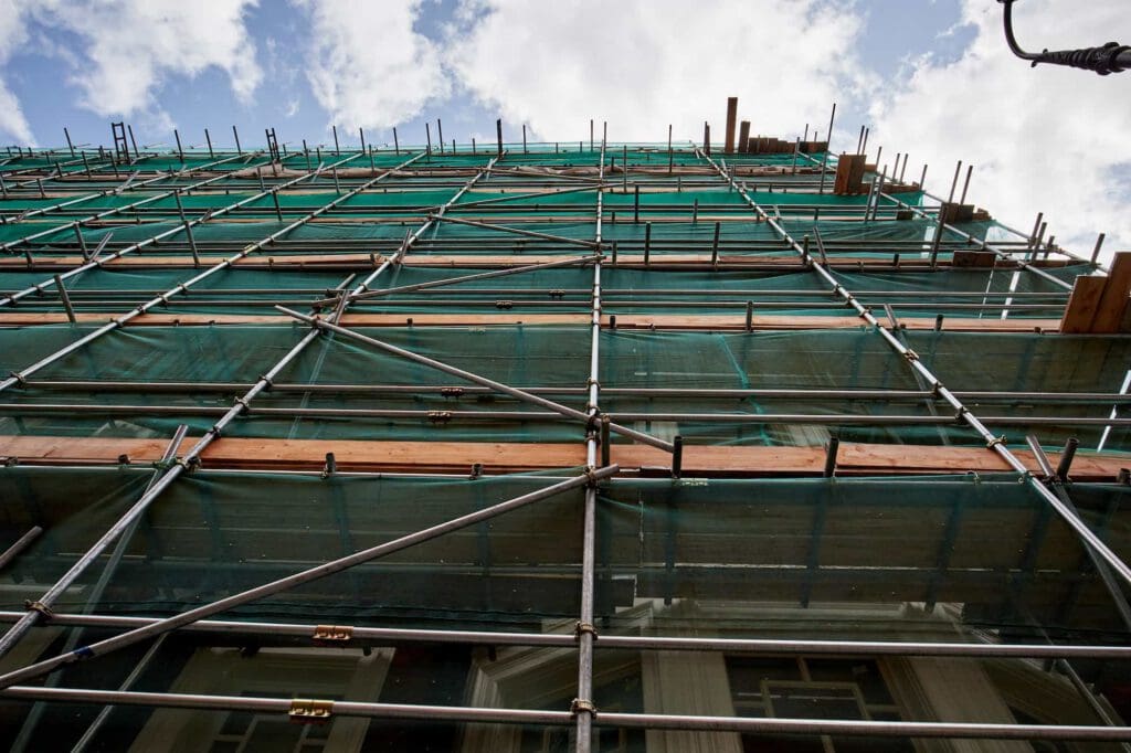 A multi-story building covered in green netting and metal scaffolding, viewed from below, with a partly cloudy sky in the background.