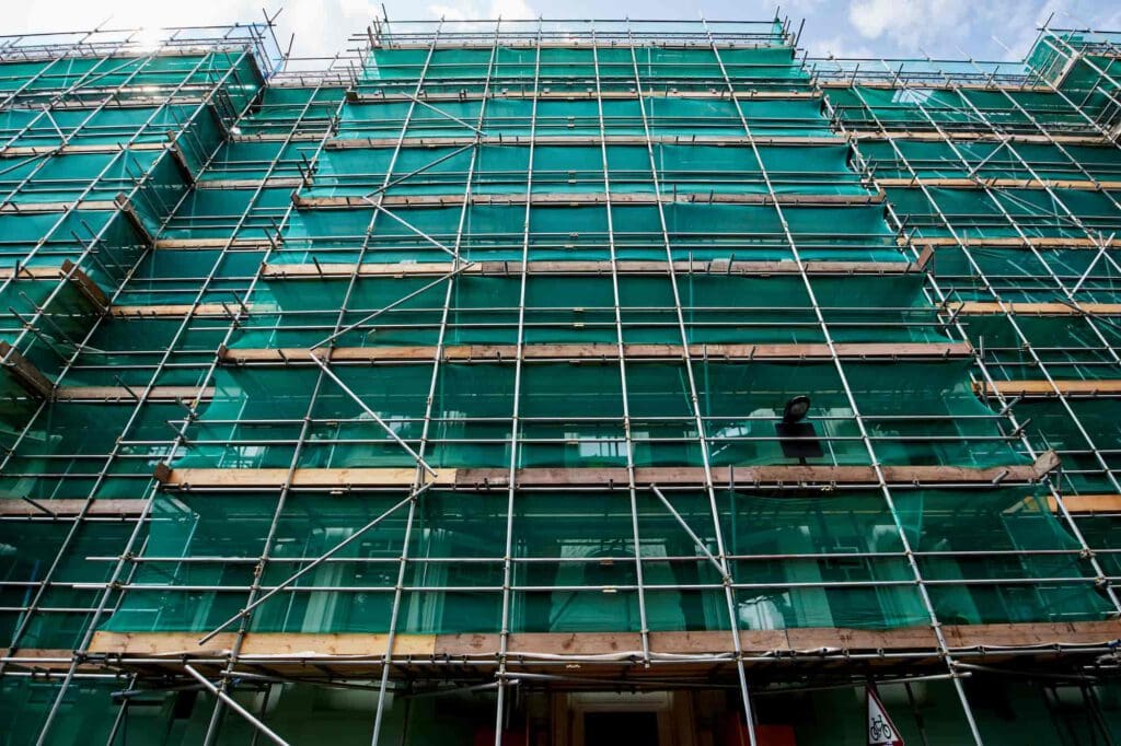 A multi-story building covered in green netting and surrounded by metal scaffolding, viewed from below against a partly cloudy sky, indicating construction or renovation work in progress.