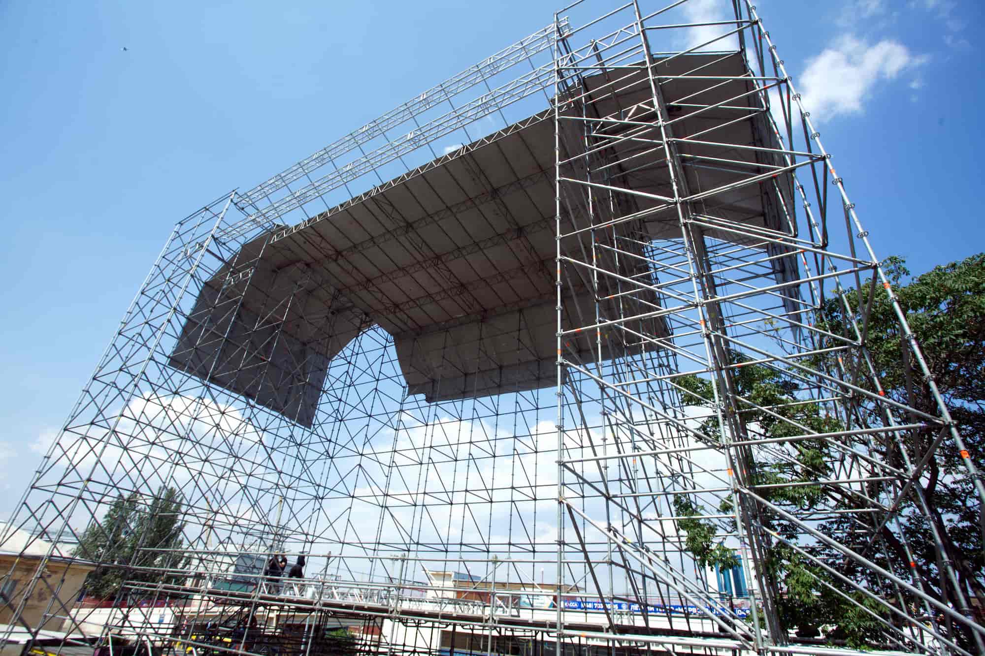 A large outdoor stage structure made of metal scaffolding is under construction against a blue sky with scattered clouds. Some trees and buildings are visible in the background.