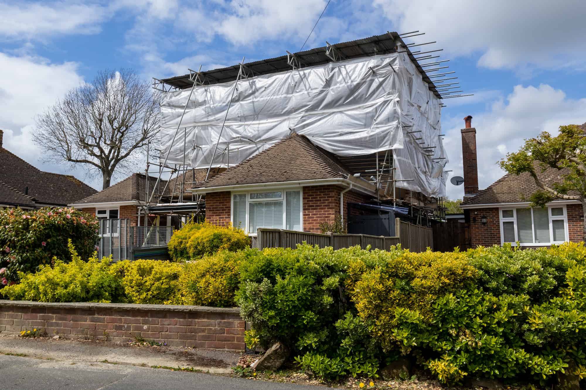 A brick house surrounded by shrubs and a low wall, with scaffolding and white tarps covering its roof and upper structure, indicating construction or renovation work. Neighboring houses and a tree are visible.