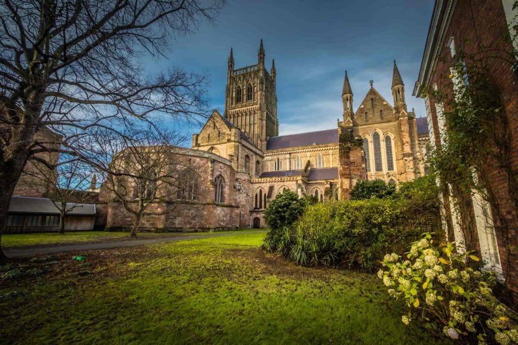 A historic stone cathedral with tall spires and arched windows stands beside a grassy lawn and garden, with trees and shrubs in the foreground under a blue sky.
