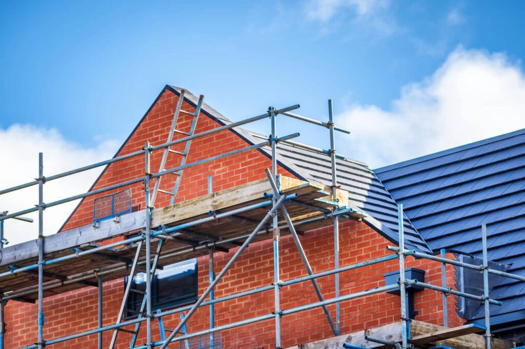 A partially constructed red brick house with scaffolding around the roof area, set against a blue sky with clouds.