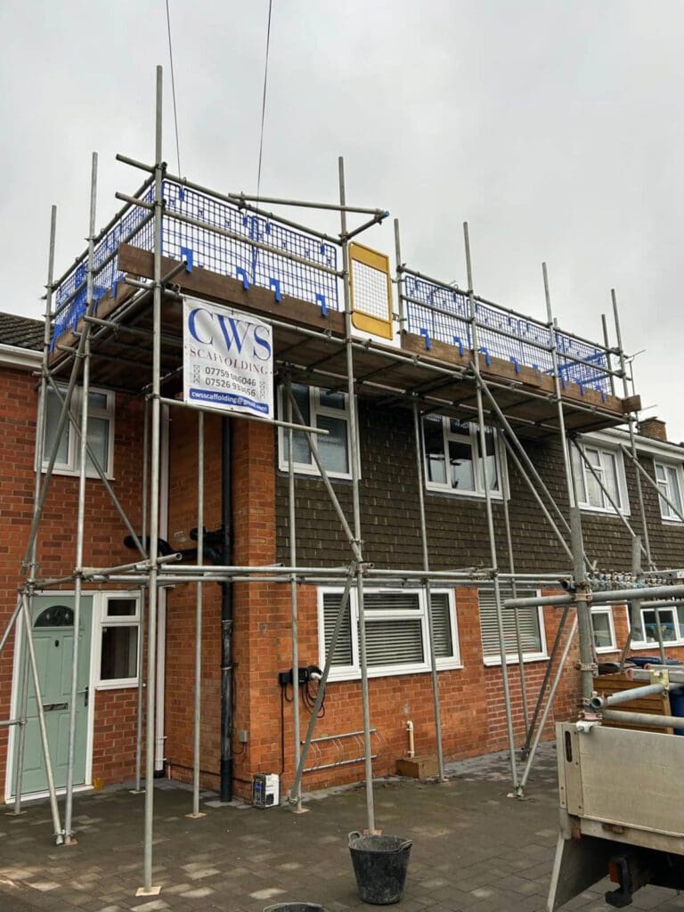 A two-story red-brick house with scaffolding set up along the front, featuring safety barriers and a sign reading "CWS Scaffolding." Construction materials and equipment are visible near the house.