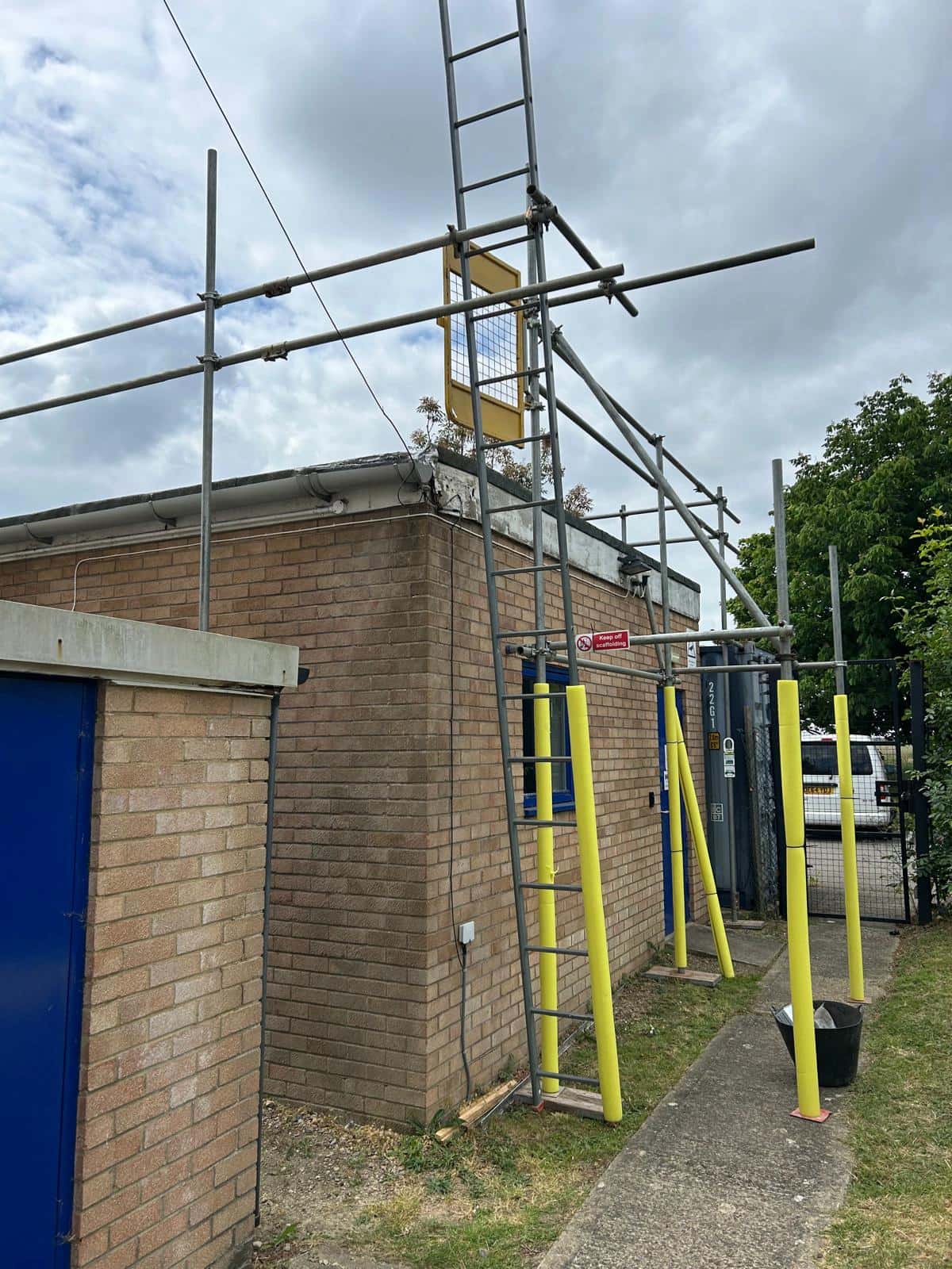 Scaffolding is set up around a brick building with a yellow ladder and guard rail. Yellow safety bollards stand at the entrance, and a cloudy sky can be seen overhead.