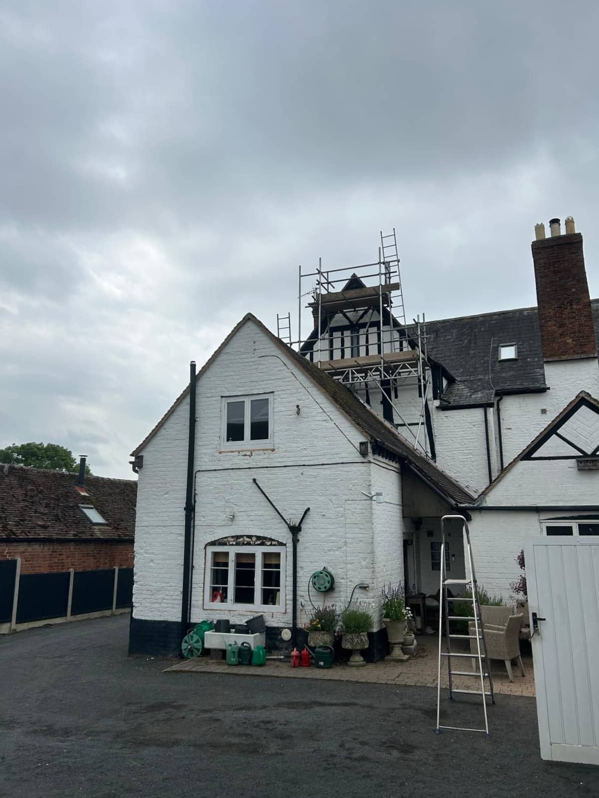 A white brick house with black trim has scaffolding set up on the roof. There’s a ladder leaning against the side, outdoor furniture, potted plants, and gardening items in the paved front area under a cloudy sky.