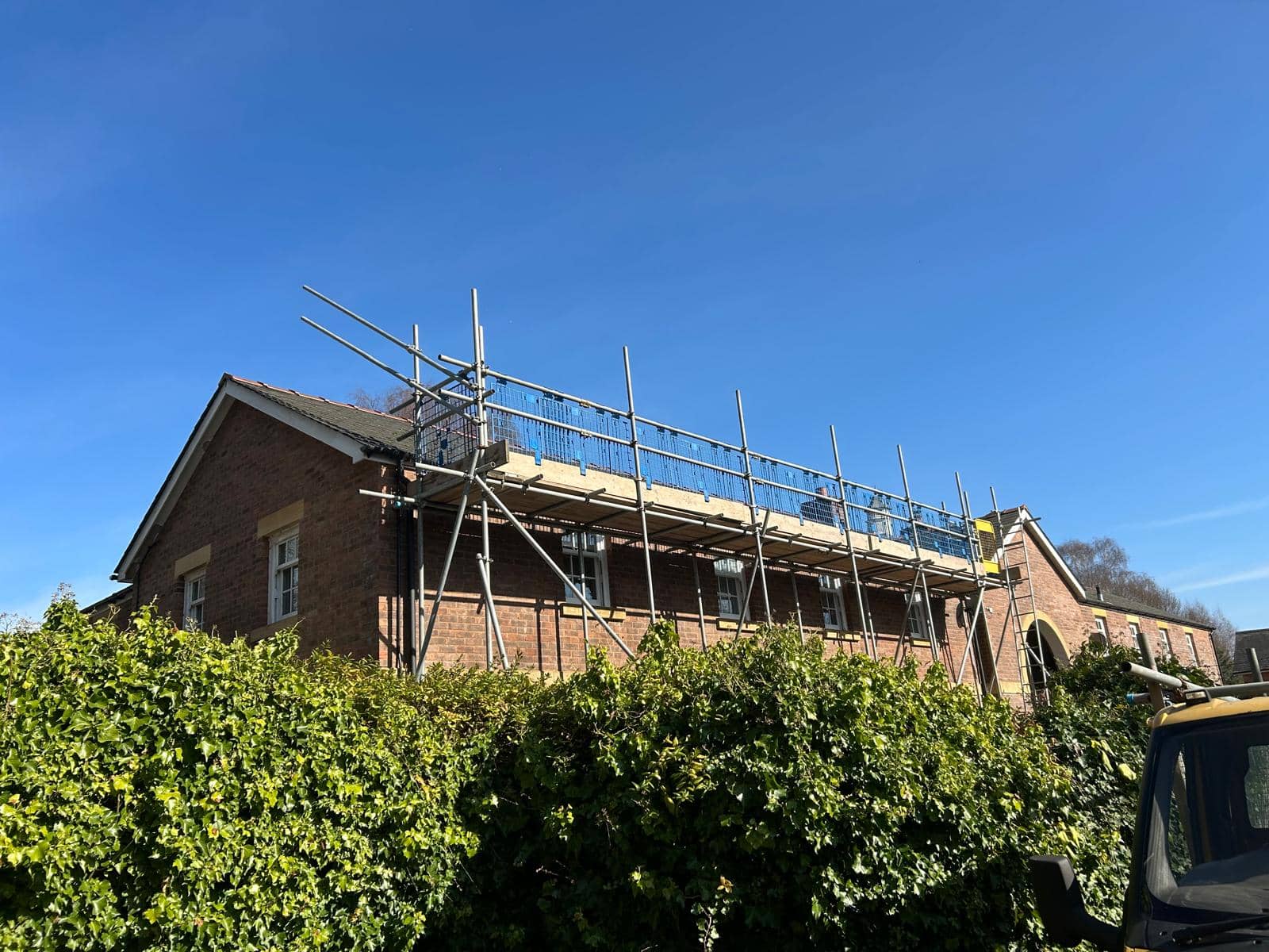 A brick building with scaffolding set up along the roofline, indicating construction or maintenance work. Green bushes line the front, and the sky is clear and blue.