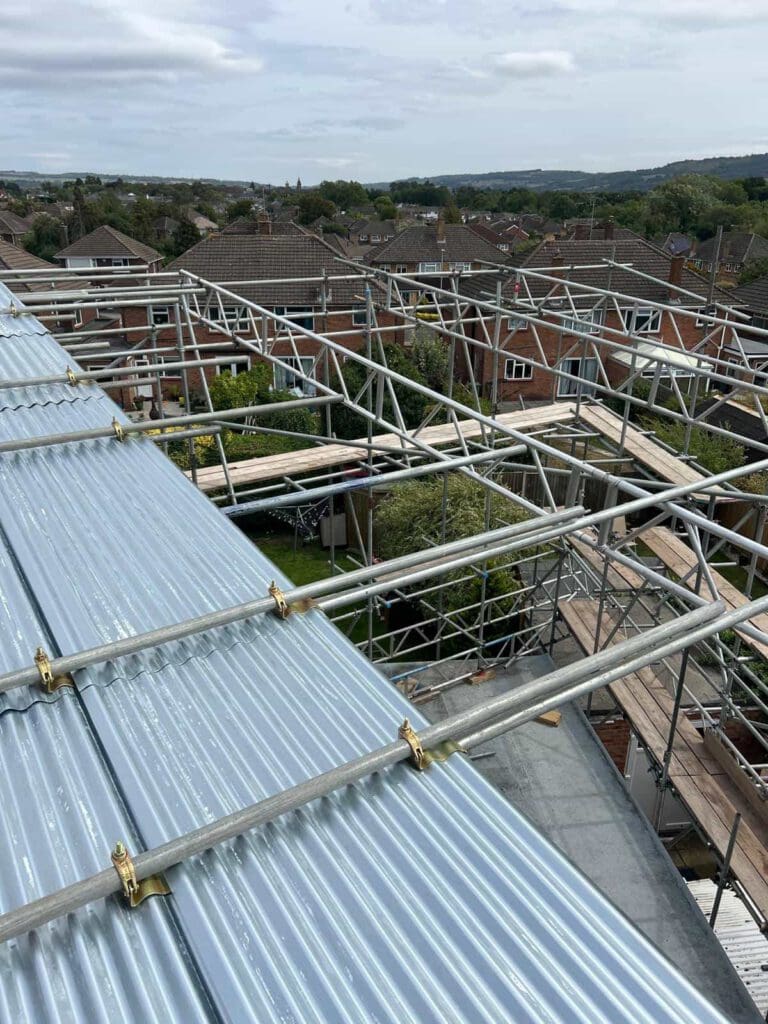 View from a rooftop showing metal roofing sheets and scaffolding structures above suburban houses, with trees and other rooftops visible in the background under a cloudy sky.