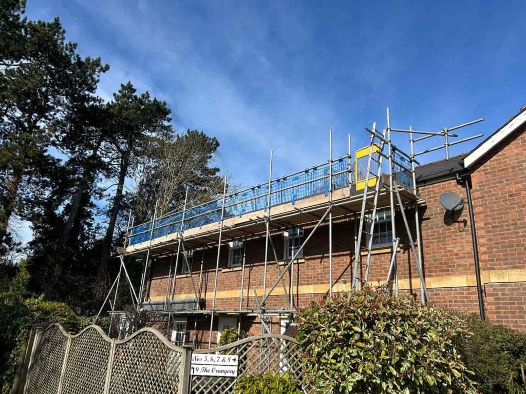 A two-story brick house with scaffolding erected along the upper level. Blue safety netting and metal poles are visible. Trees and bushes surround the house, and a sign hangs on the fence in front. The sky is clear and blue.