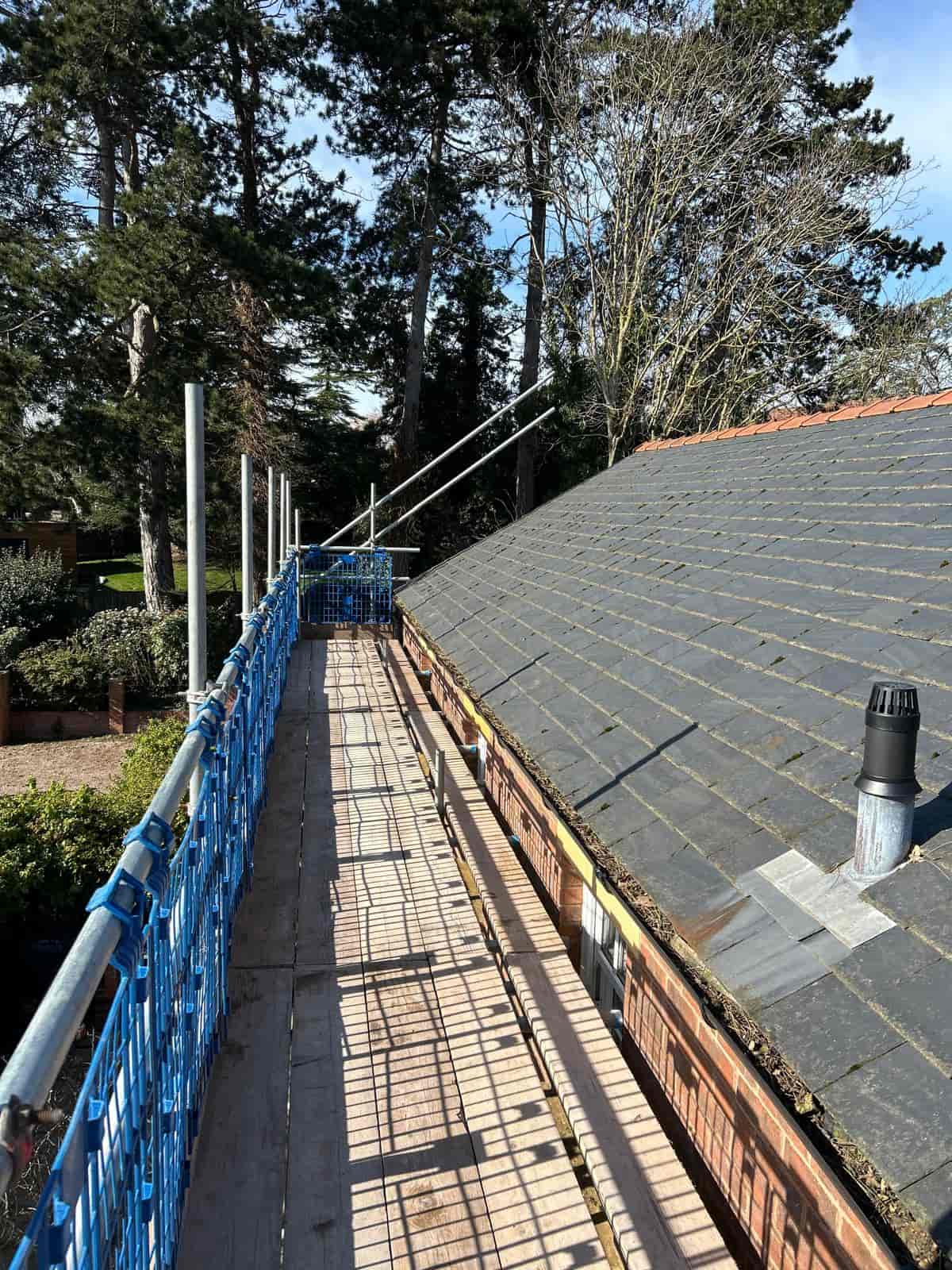 A view of a rooftop with gray slates next to a wooden scaffolding platform secured with blue safety barriers, surrounded by tall trees under a clear sky.