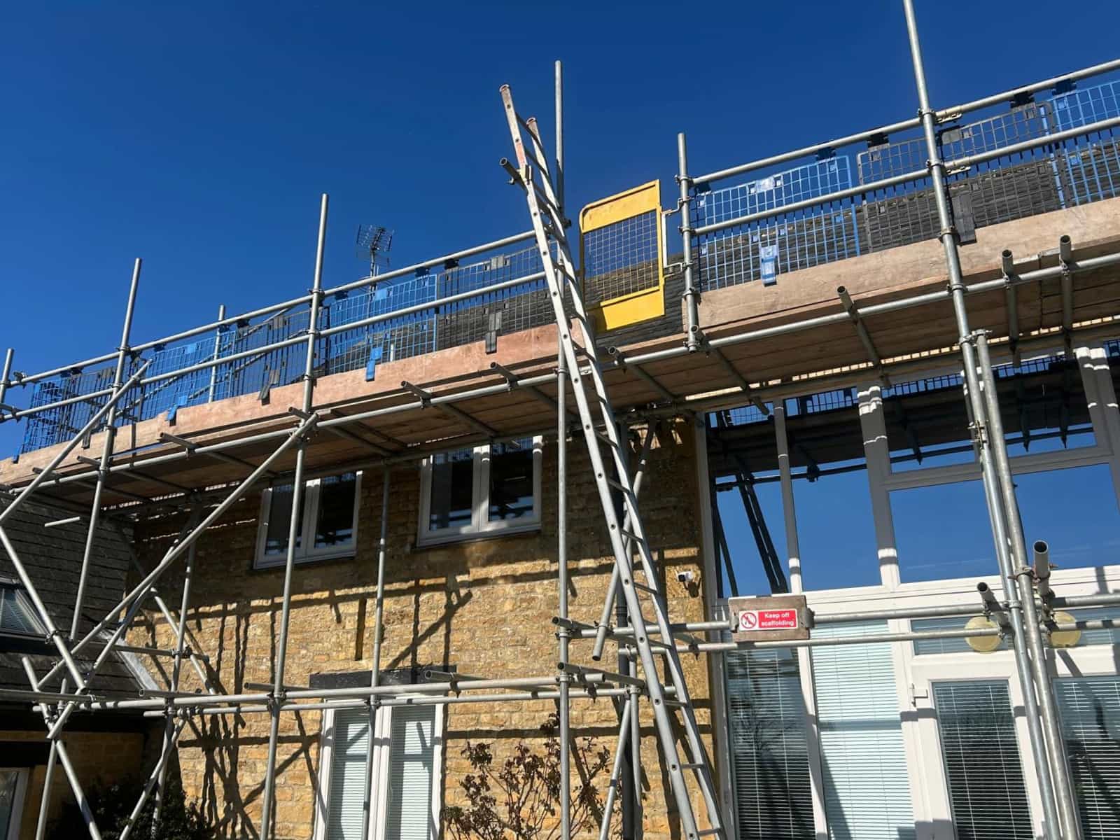 Scaffolding is set up around a two-story brick house with large windows and a glass conservatory, under a clear blue sky.