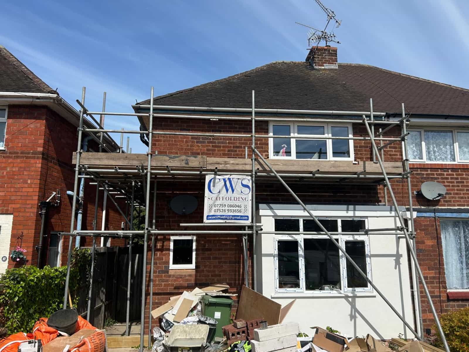 A two-story brick house with scaffolding on the front, a construction company sign, and debris on the ground, indicating ongoing renovation or building work.