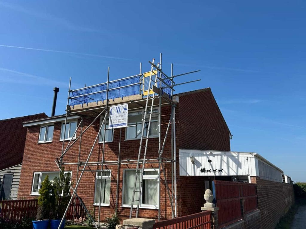 A brick semi-detached house with scaffolding erected around the chimney for construction work, under a clear blue sky. A sign on the scaffolding reads "CWS.