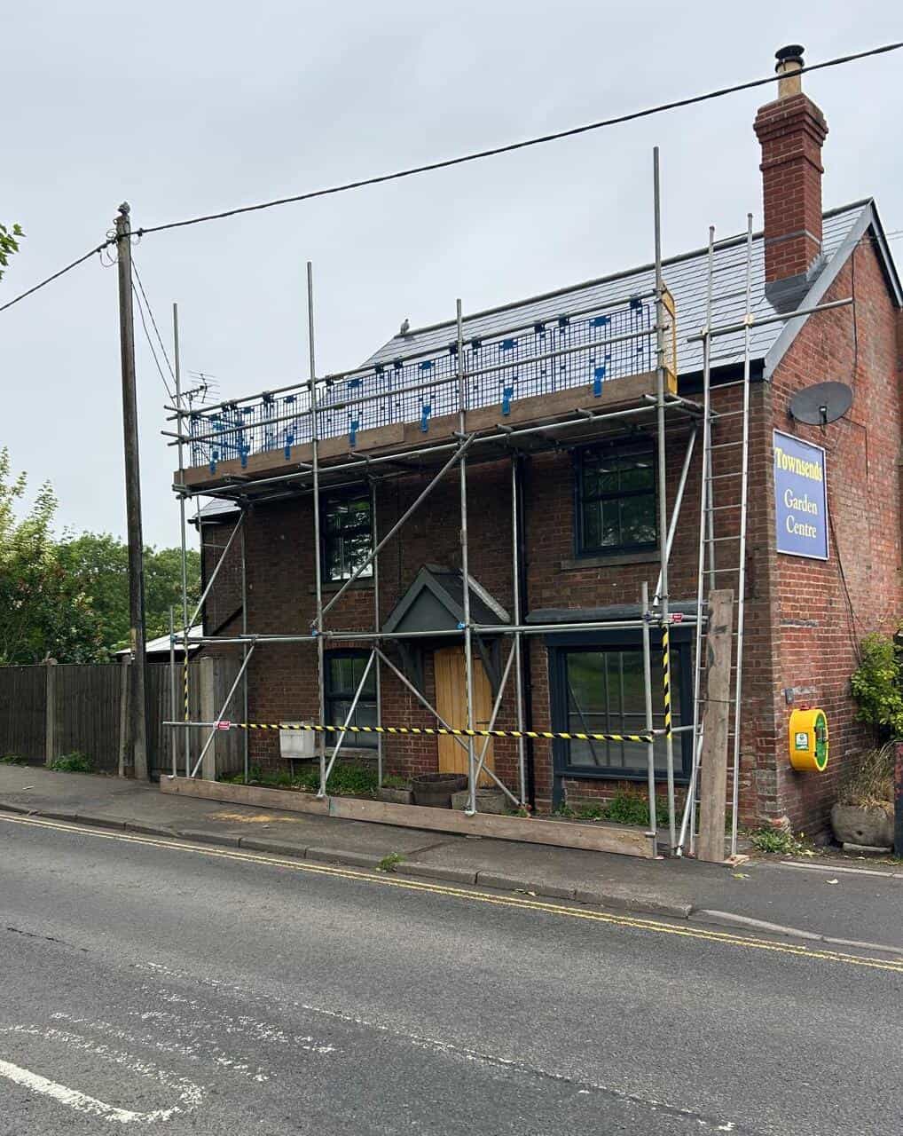 A brick building with scaffolding covering the front and roof, cordoned off with caution tape. A sign reads "Townsend Garden Centre." The building sits beside a road, with an emergency defibrillator mounted on the wall.
