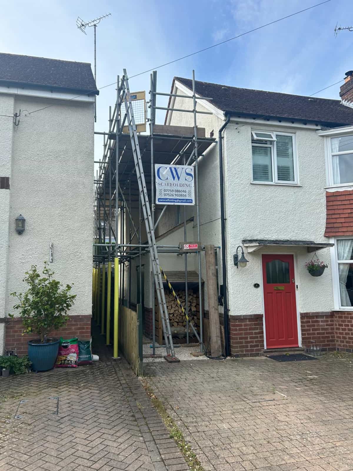 Scaffolding is set up between two white houses, with a sign reading “CWS Scaffolding” attached. A woodpile is stored beneath the scaffolding, and one house has a red front door. The driveway and some potted plants are visible.