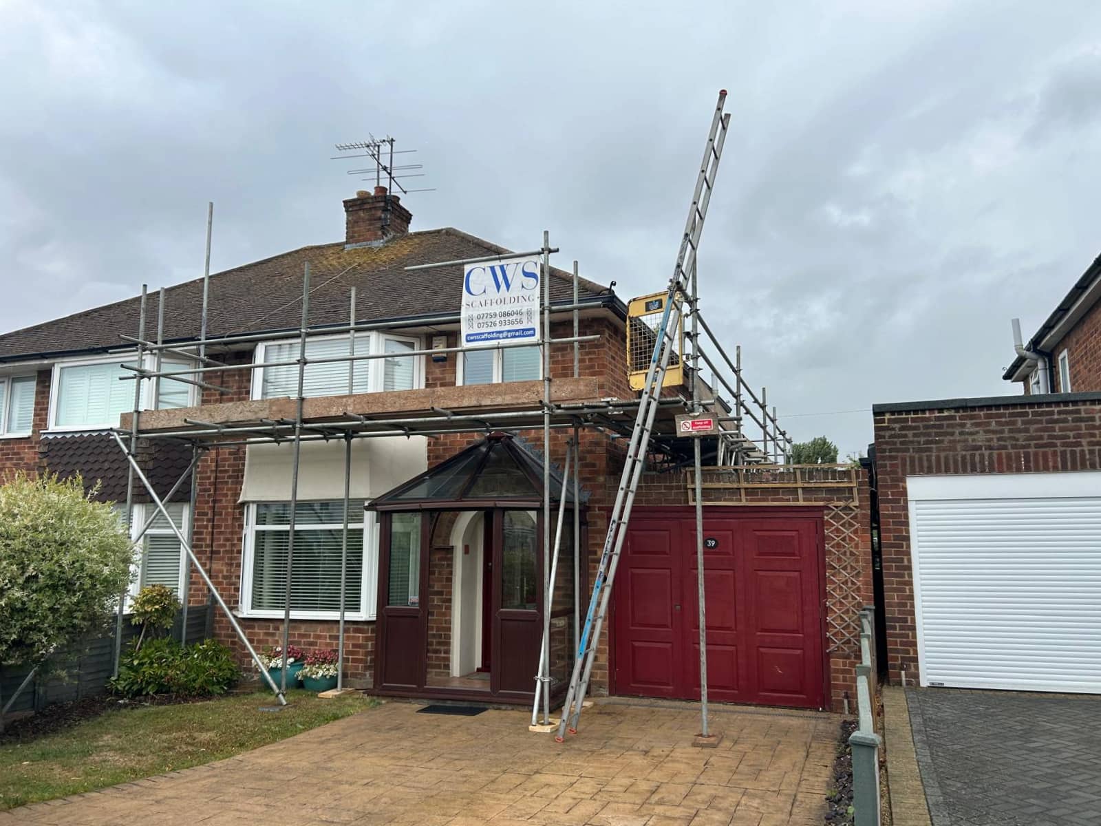 A brick semi-detached house with scaffolding and ladders set up around the roof, a red garage door, a sign reading “CWS” on the scaffolding, and a cloudy sky overhead.