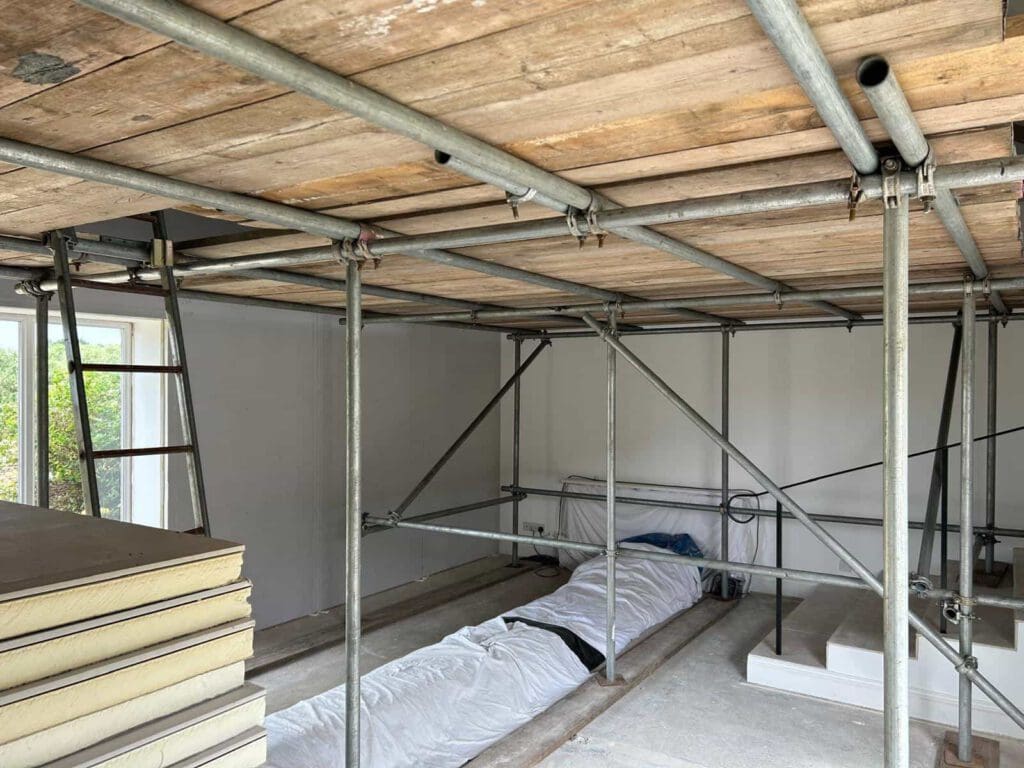 Indoor construction site with metal scaffolding and a wooden platform overhead, stacked drywall sheets on the left, and a covered object on the floor in the center. White walls and a window are visible in the background.