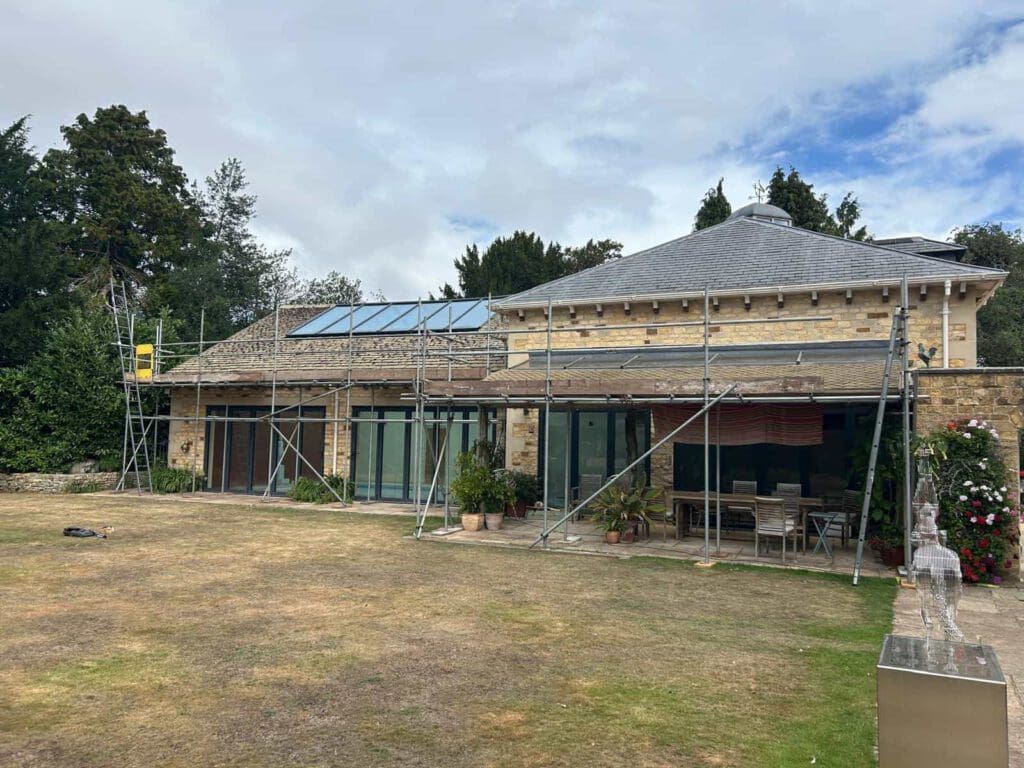 A stone house with large glass doors is surrounded by scaffolding on the lawn, indicating construction or renovation work. Potted plants and outdoor furniture are visible under a covered area.