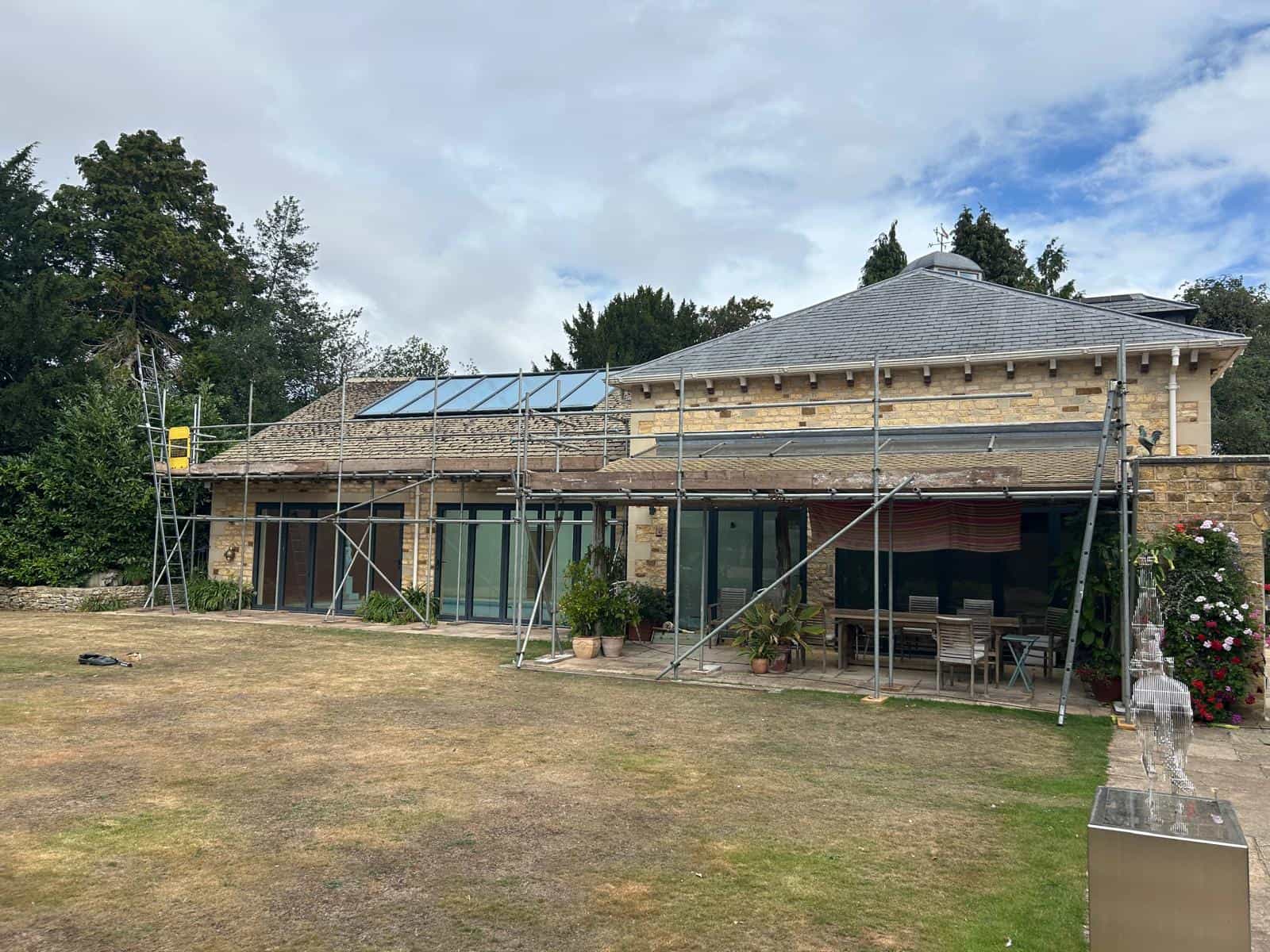 A stone house with large glass doors is surrounded by scaffolding on the lawn, indicating construction or renovation work. Potted plants and outdoor furniture are visible under a covered area.
