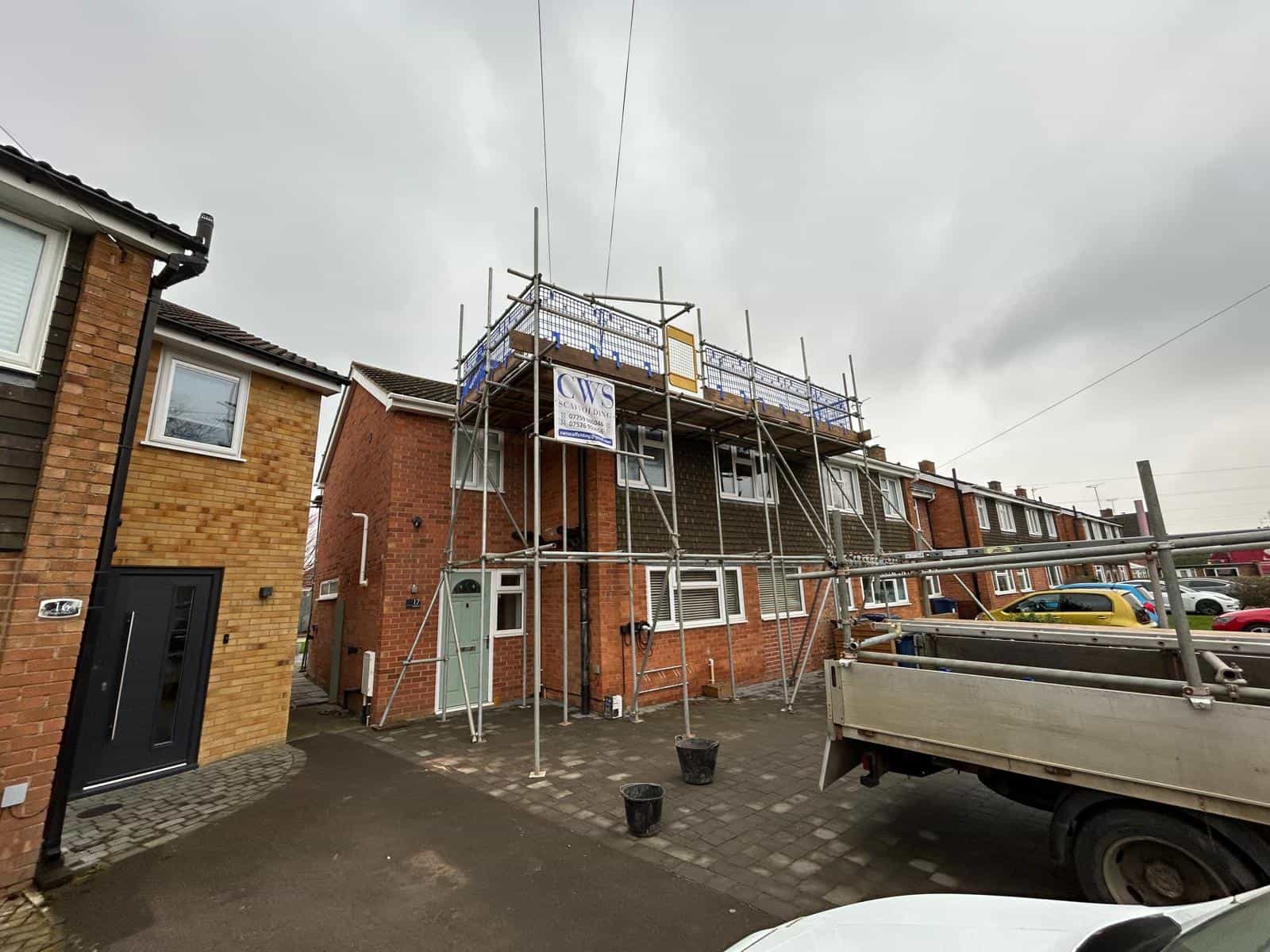 A two-story brick house with scaffolding set up on the front for construction or repairs. A sign and safety barriers are attached to the scaffolding. A truck is parked nearby on a cloudy day.