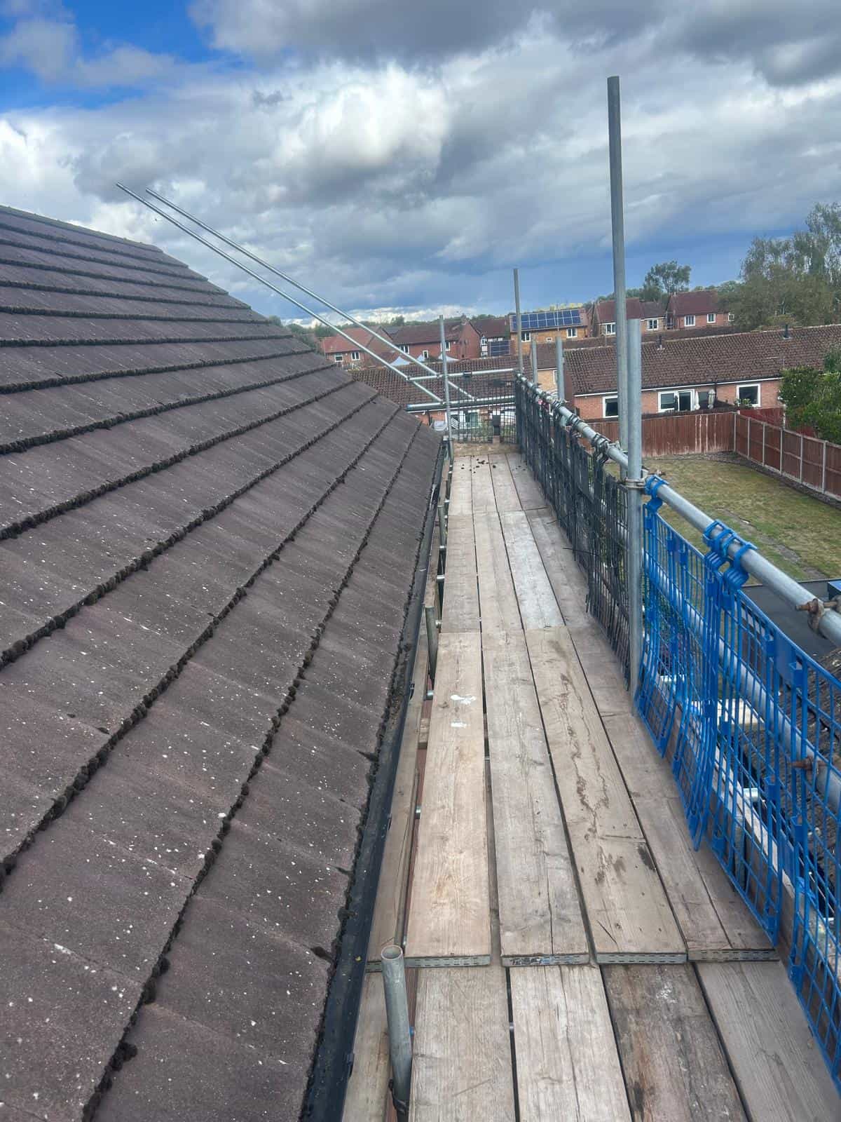 A wooden scaffolding walkway runs alongside a sloped roof with brown shingles. Blue safety netting and metal poles are on the right, overlooking neighboring houses under a cloudy sky.