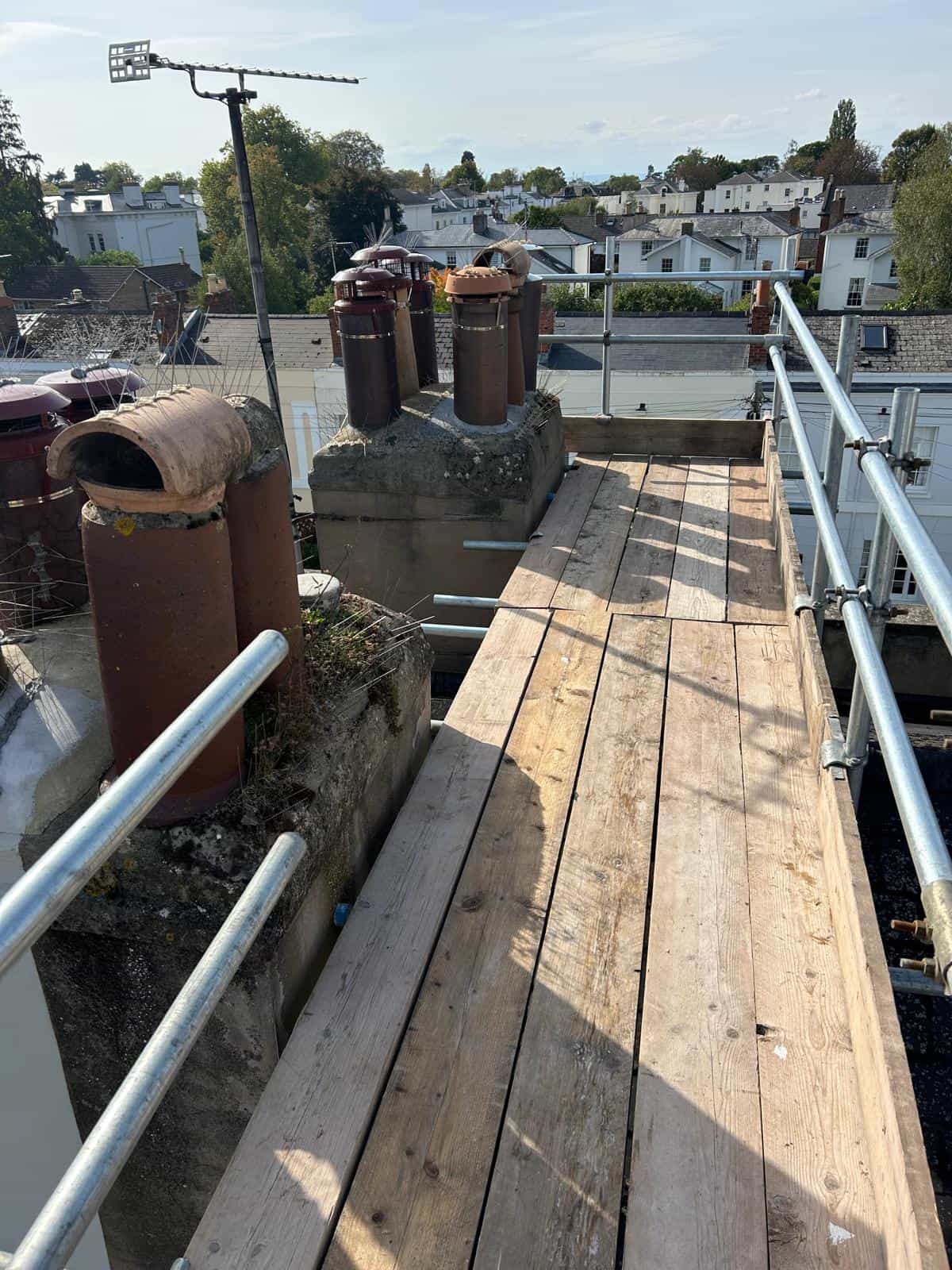 A wooden scaffold walkway with metal railings runs alongside rooftop chimneys and vents, overlooking residential houses and trees in the background on a sunny day.