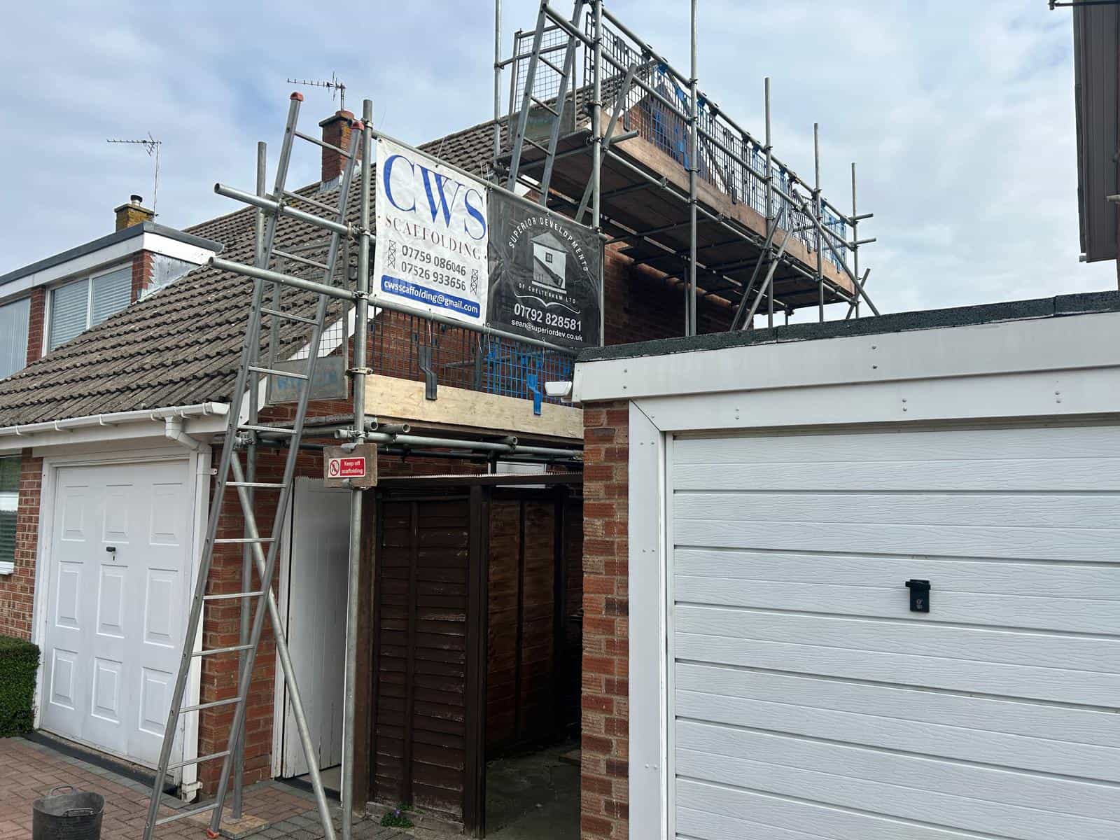 A house under construction with scaffolding set up above the garage. Two business signs are attached to the scaffolding. The white garage door is closed, and the sky is partly cloudy.