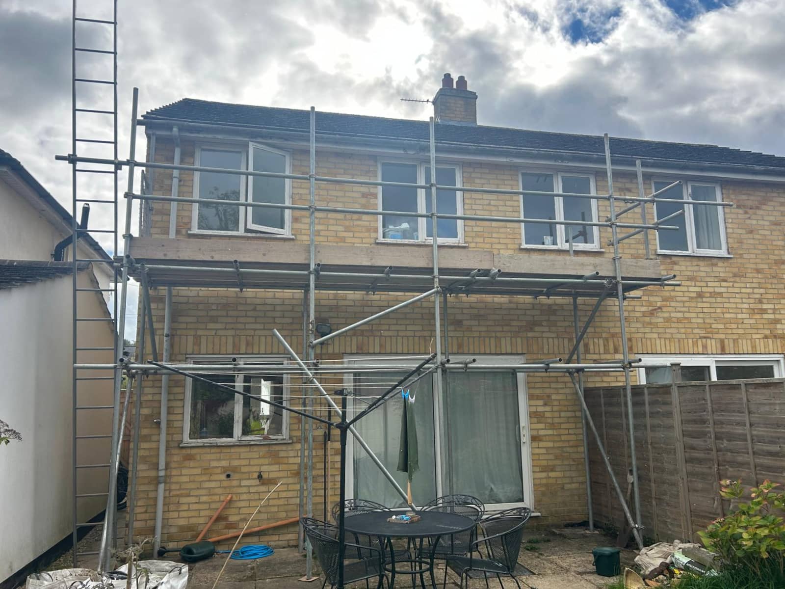 A two-story brick house with scaffolding set up along the front. The patio below has a round table and chairs, and some tools and garden items are scattered nearby. The sky is partly cloudy.