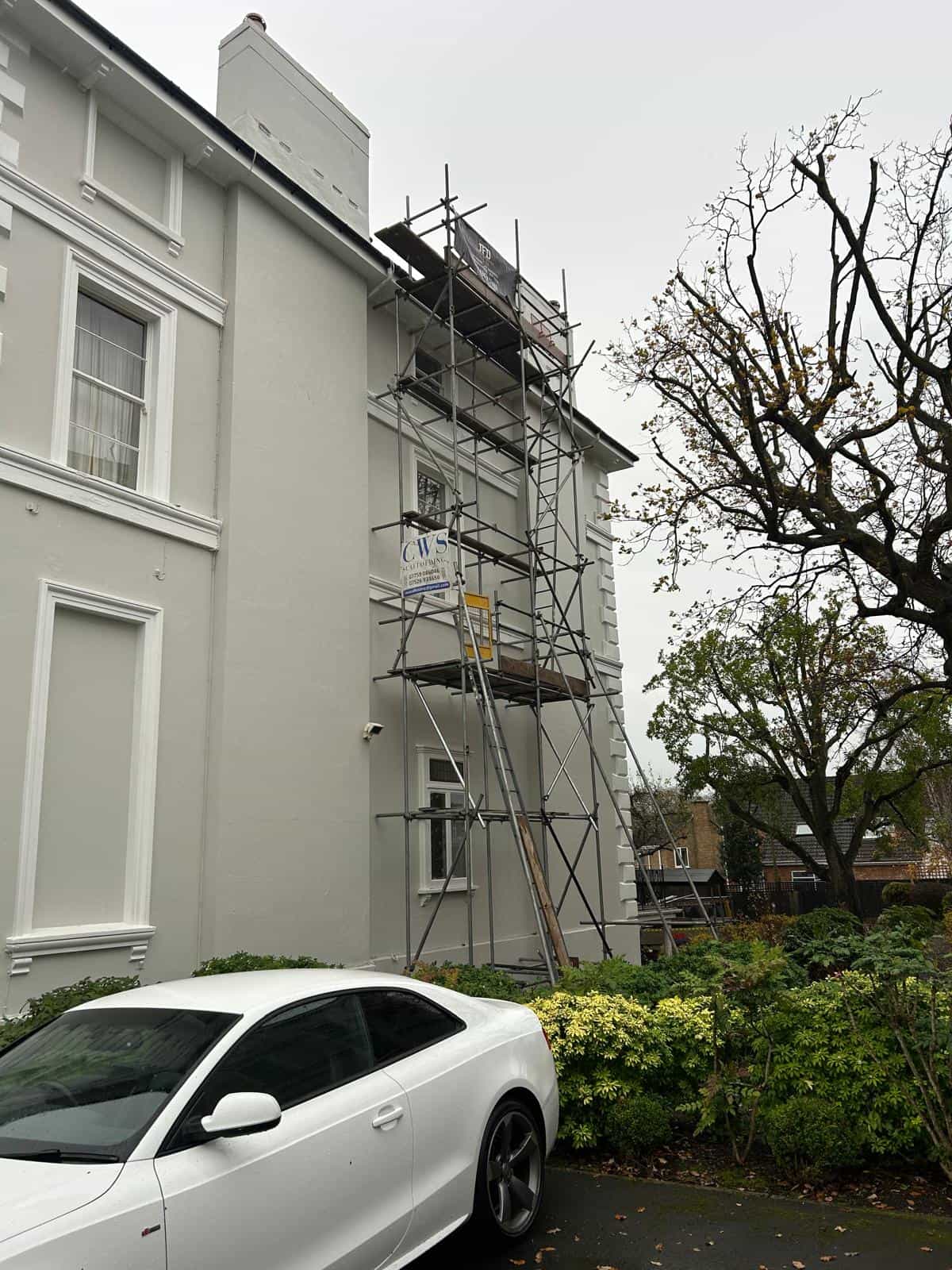 A white car is parked in front of a light grey building with scaffolding set up against its side. Some trees and bushes are nearby, and the sky is overcast.