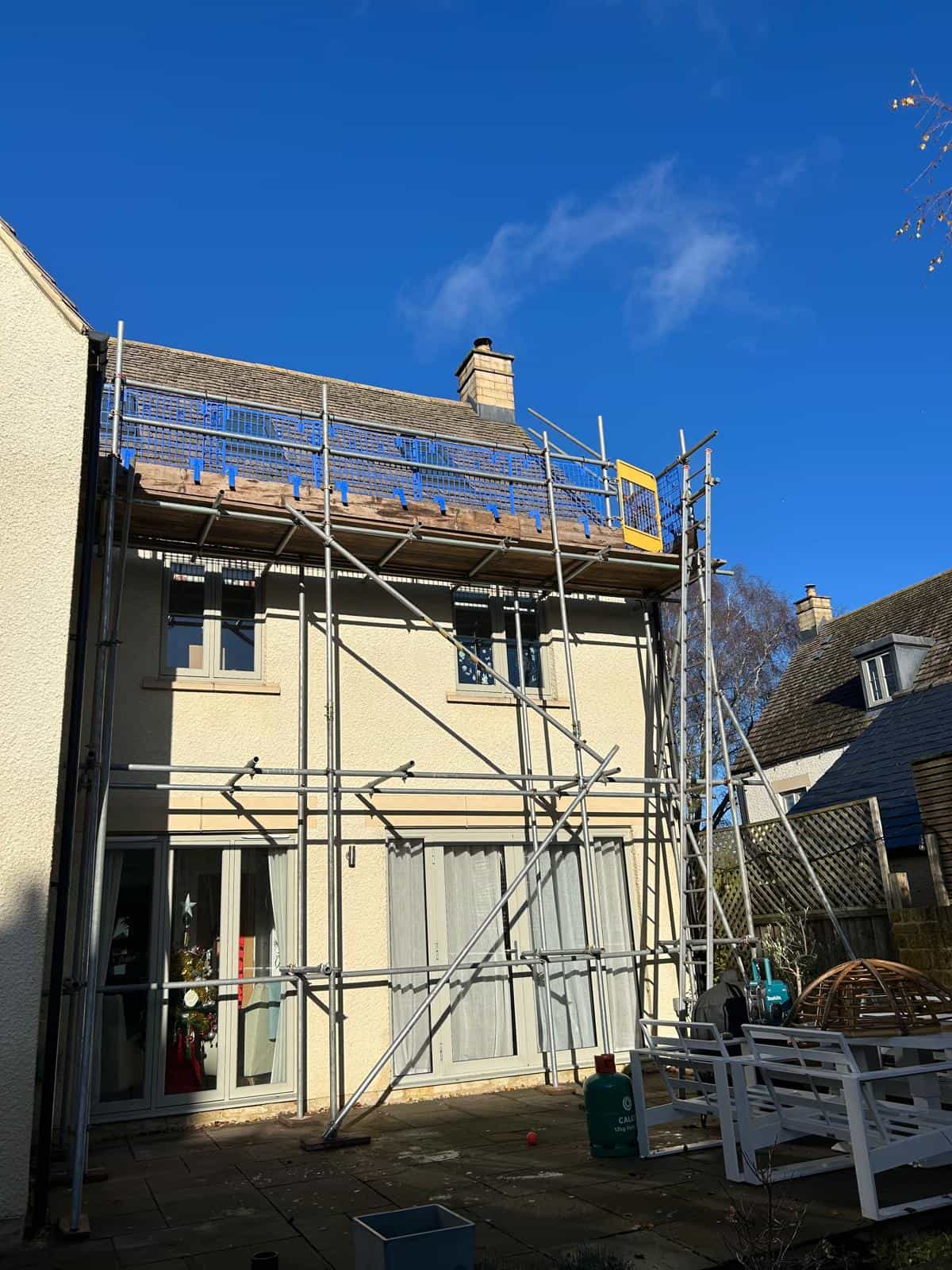 A two-story house with scaffolding set up along the front, likely for roof or exterior work. The sky is clear and blue, and the patio below has various outdoor items and furniture.