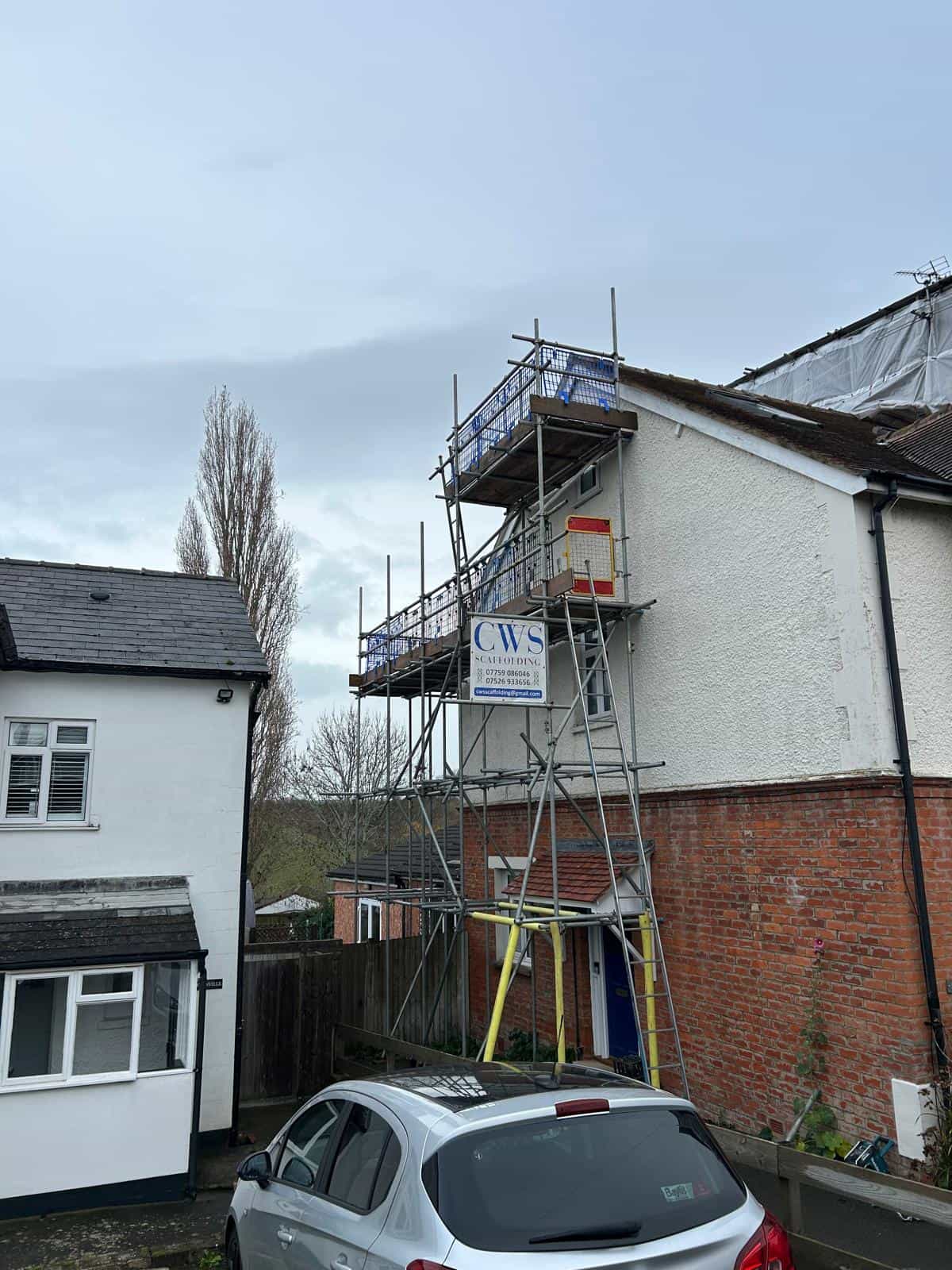 A house with scaffolding set up against one side, displaying a CWS Construction sign. A gray car is parked in front, and the sky is overcast. Another house is partially visible on the left side of the image.