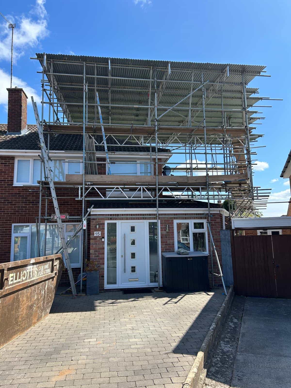 A brick semi-detached house with extensive metal scaffolding and a corrugated roof covering the top, likely for construction or roof work, under a clear blue sky. A skip is on the left of the driveway.