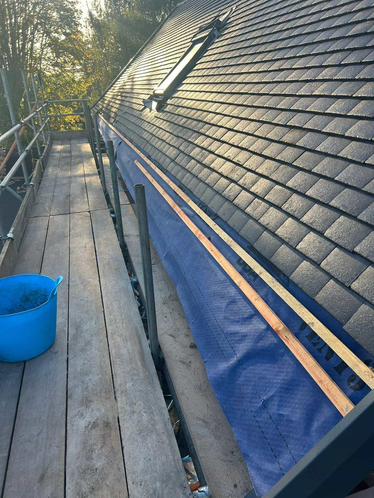 A rooftop under construction with scaffolding along the edge, a blue tarp partially covering the roofline, wooden planks on the scaffolding, and a blue bucket nearby; sunlight shines on the slate roof tiles.