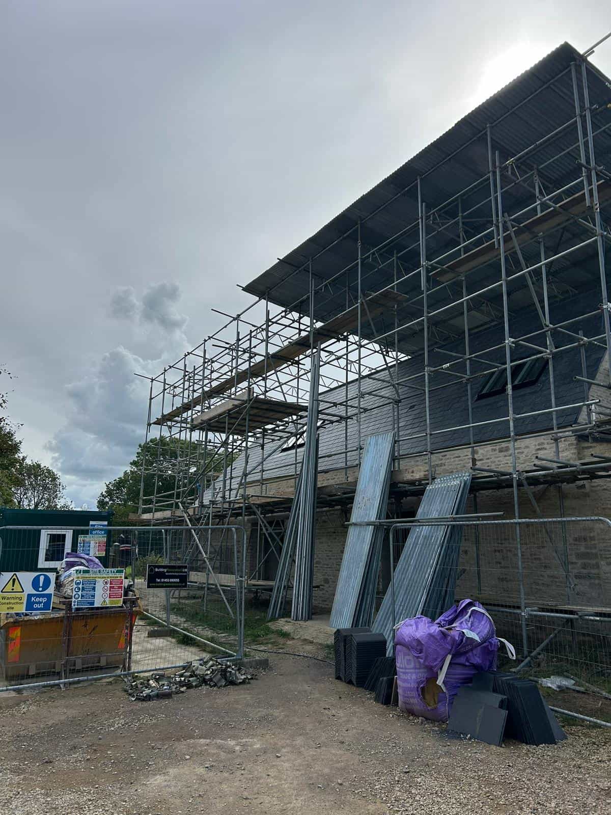 A building under construction, covered in scaffolding, with metal sheets, a purple bag, and construction materials nearby. A dumpster, warning signs, and a cloudy sky are also visible.