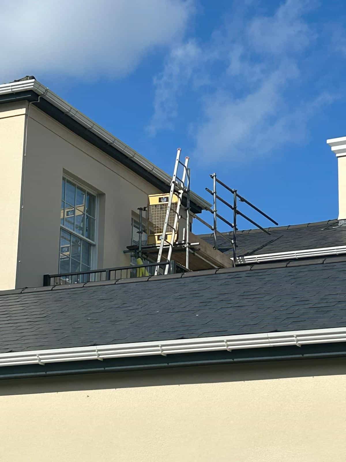 A tall ladder leans against the side of a cream-colored building, reaching up to a balcony with metal railings and construction materials. The sky is bright blue with a few clouds.