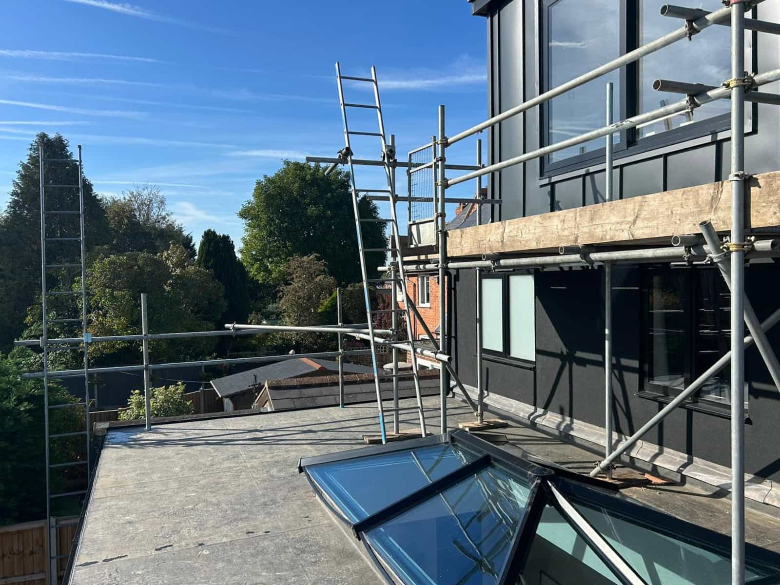 Scaffolding is set up along the edge of a flat roof next to a modern building with large windows. Trees and houses are visible in the background under a clear blue sky.