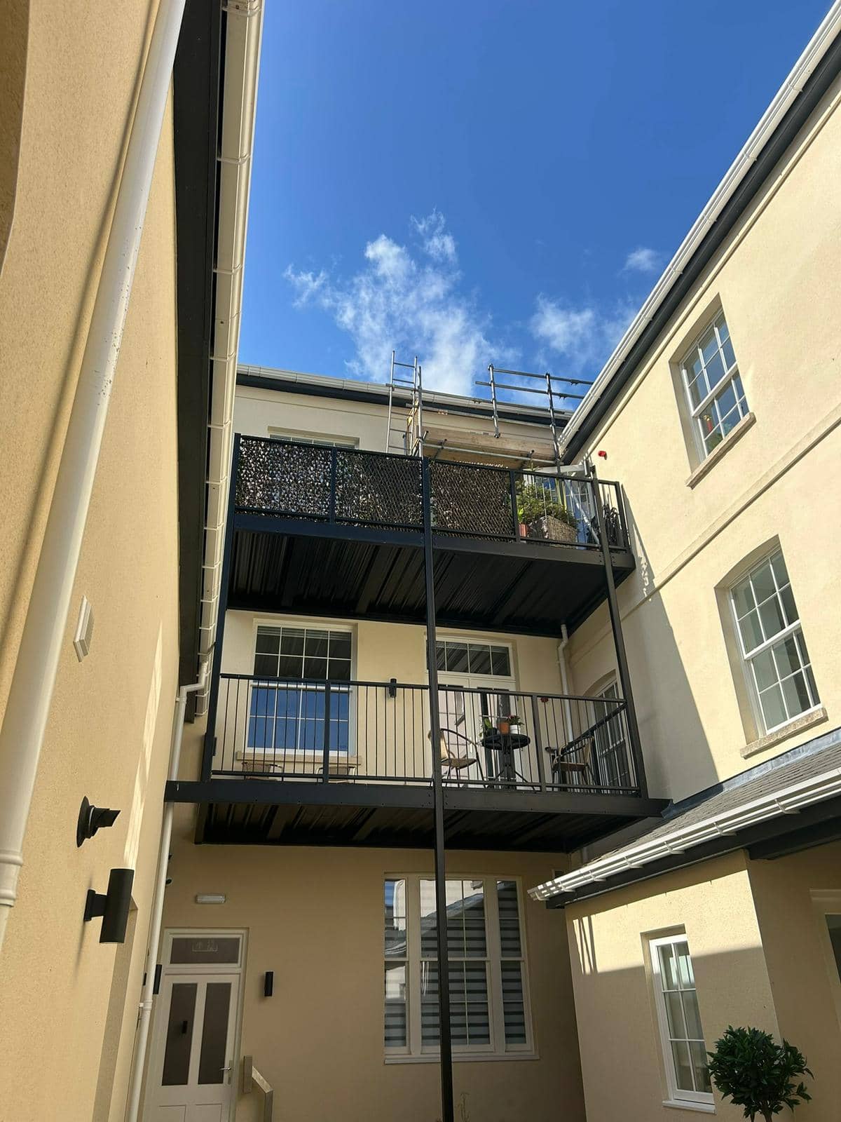 A view looking up at the exterior of a light-colored apartment building with balconies, black railings, and a partly cloudy blue sky above. There are chairs and plants on the middle balcony.