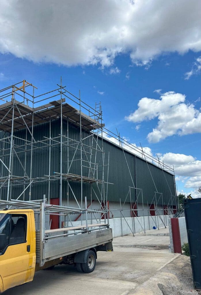 A large industrial building with scaffolding around its dark green metal exterior. A yellow and silver flatbed truck is parked in front. The sky is blue with scattered white clouds.