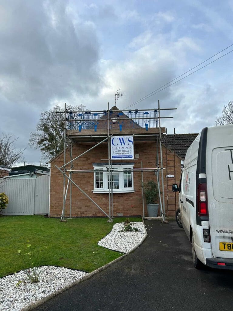 A brick house with scaffolding set up in front, a sign reading "CWS" attached to the scaffolding, and a white van parked in the driveway. The sky is cloudy and there are plants beside the driveway.