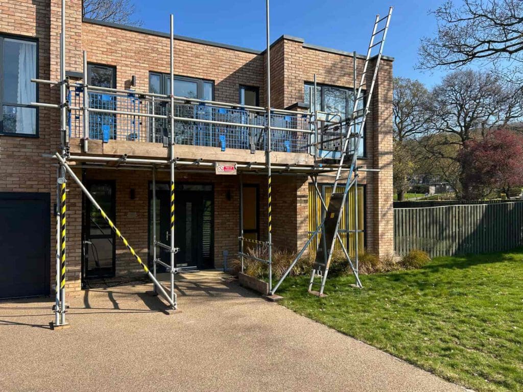 Two-story brick house with scaffolding and a ladder set up around the front balcony for construction or repairs. The yard is grassy, and the sky is clear and sunny.